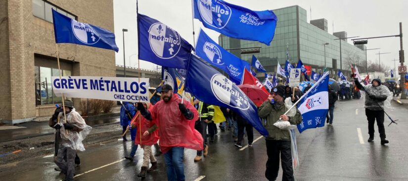 A parade of striking Steelworkers marching down a street waving, waving Métallos flags.